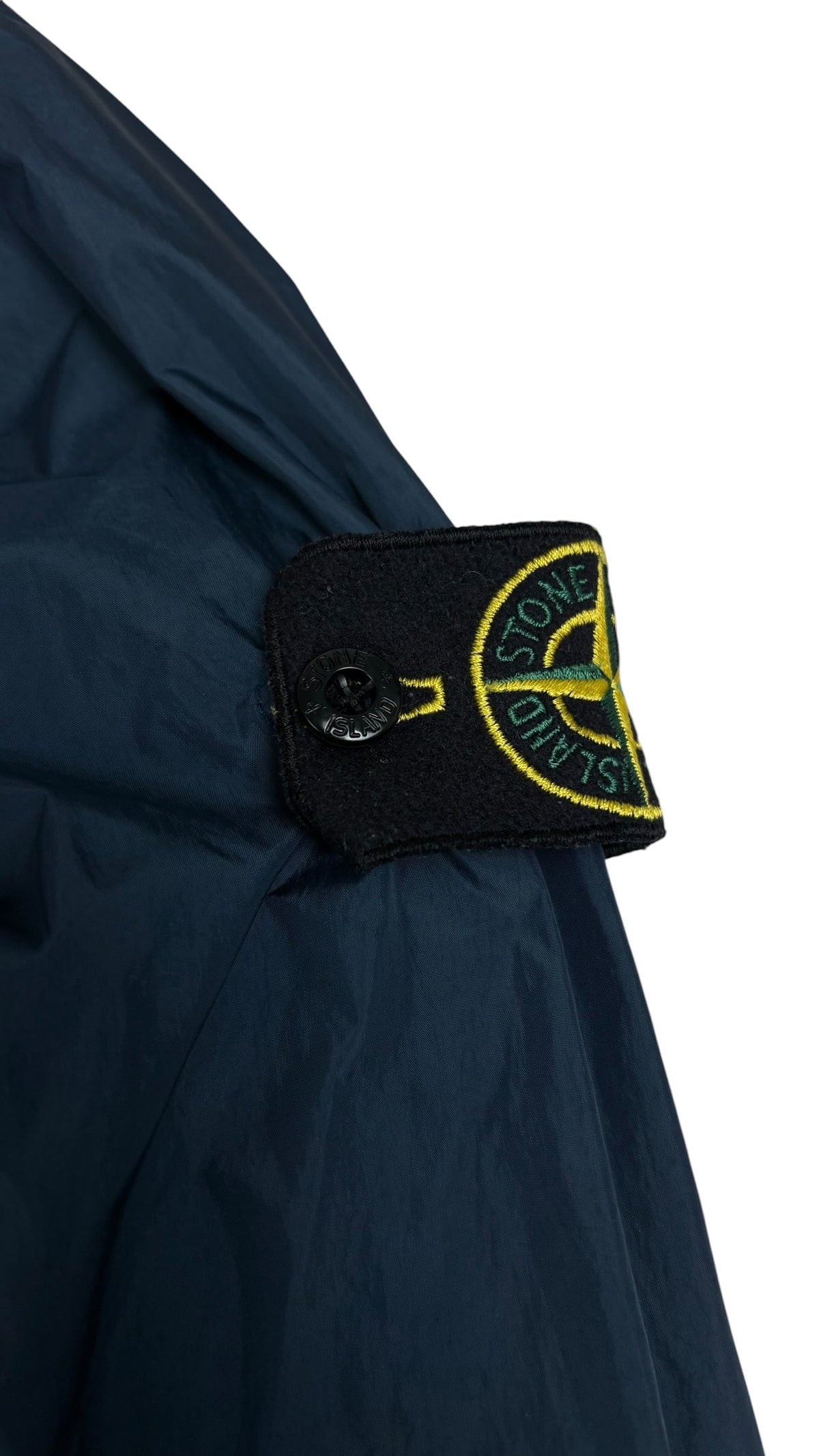 Gros plan sur l'écusson boussole Stone Island jaune et vert, badge amovible fixé par boutons sur la manche de la veste coupe-vent bleu marine, vue détail