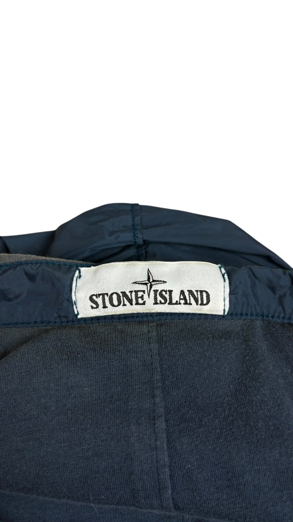 Gros plan sur l'étiquette de marque Stone Island tissée blanche avec le logo boussole, cousue à l'intérieur de la veste coupe-vent bleu marine, vue détail